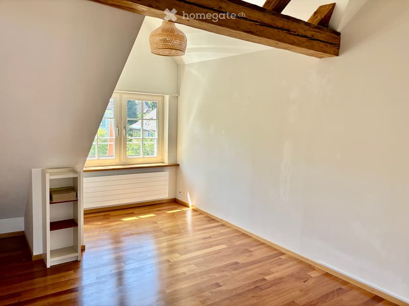 An attic room with wooden floors, white walls, exposed wooden beams, a window with a view, and a small built-in shelf unit.