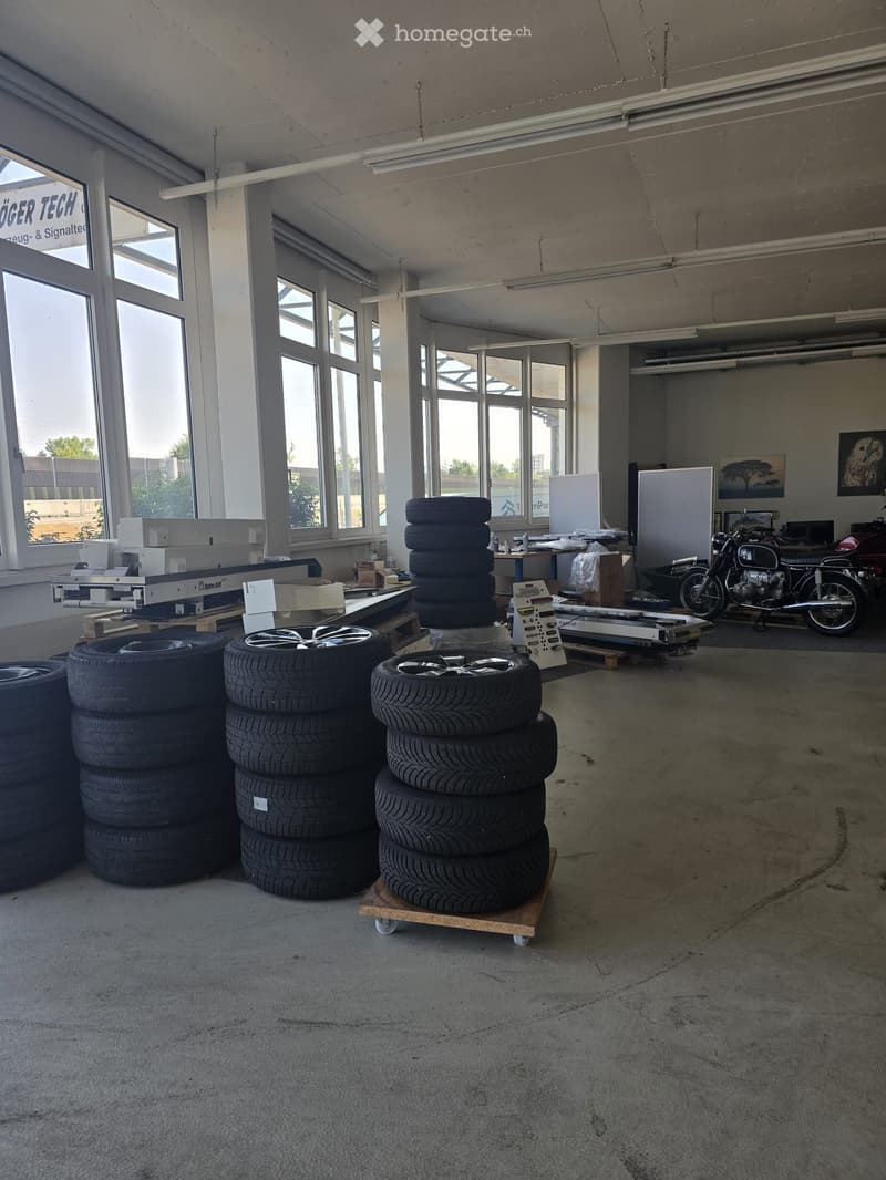 Large industrial-style room with concrete floors, large windows, stacks of tires, a motorcycle, a table, and artwork on the walls.