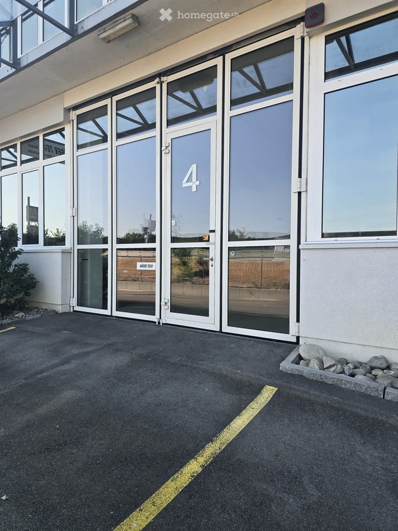 Exterior view of a modern building with large glass doors marked with the number 4, a small landscaped area with rocks, and a paved ground with a yellow line.