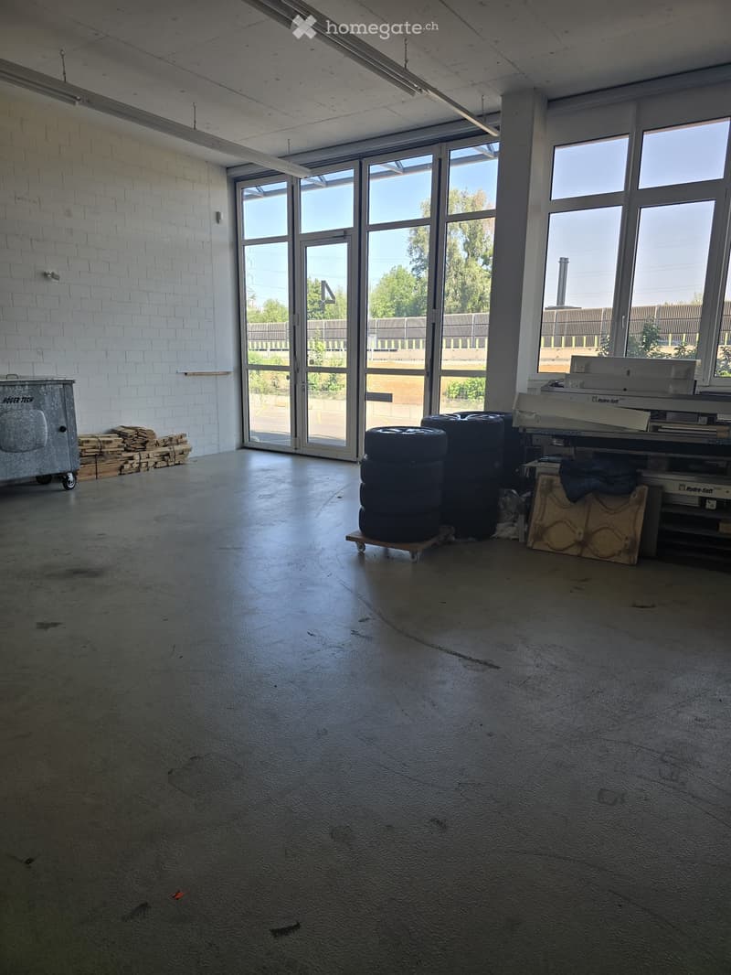 Empty industrial-style room with concrete floors, white brick walls, large windows, and a few scattered items including tires, a metal cart, and a stack of wooden planks.