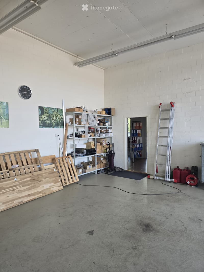 A spacious, unfinished garage with concrete flooring, white brick walls, and exposed metal beams. Contains shelves with boxes, a ladder, a vacuum cleaner, and wooden planks.
