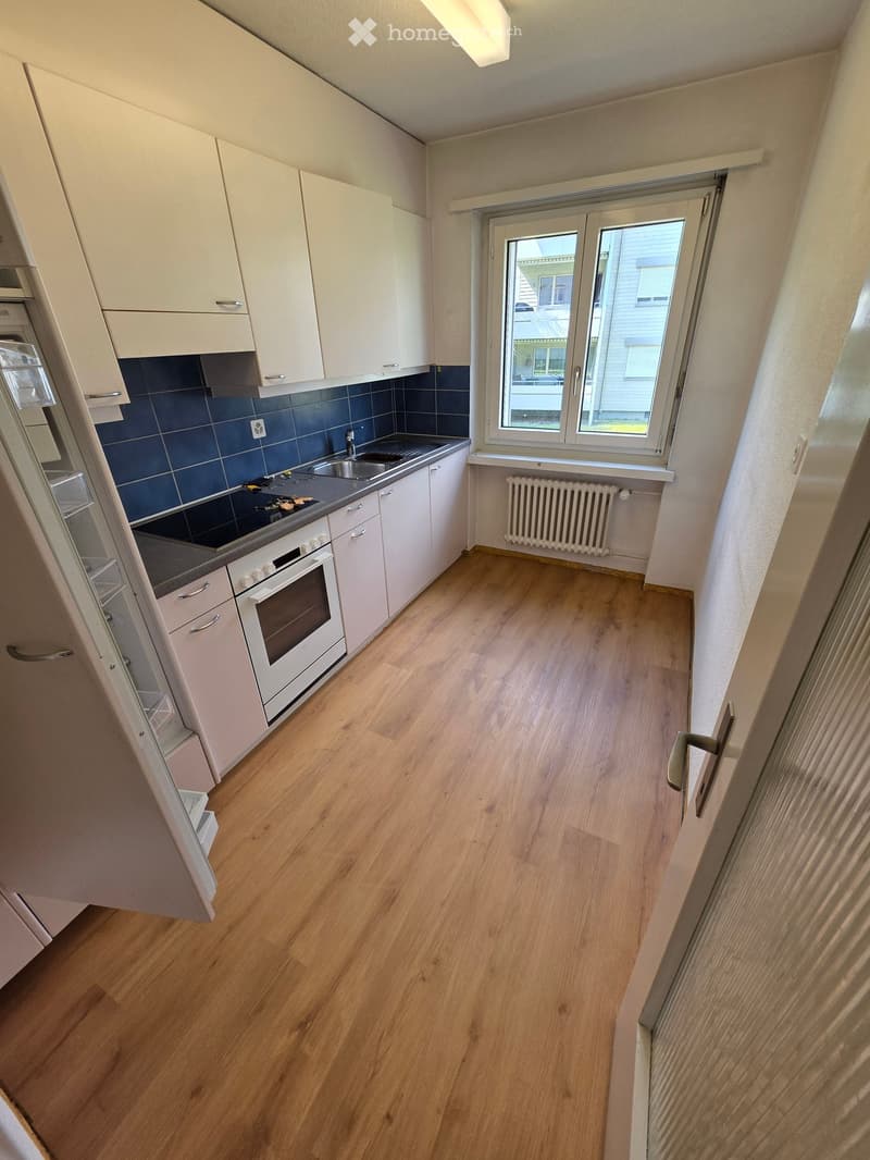 Modern kitchen with light wooden flooring, white cabinets, stainless steel appliances, and a window overlooking the street.