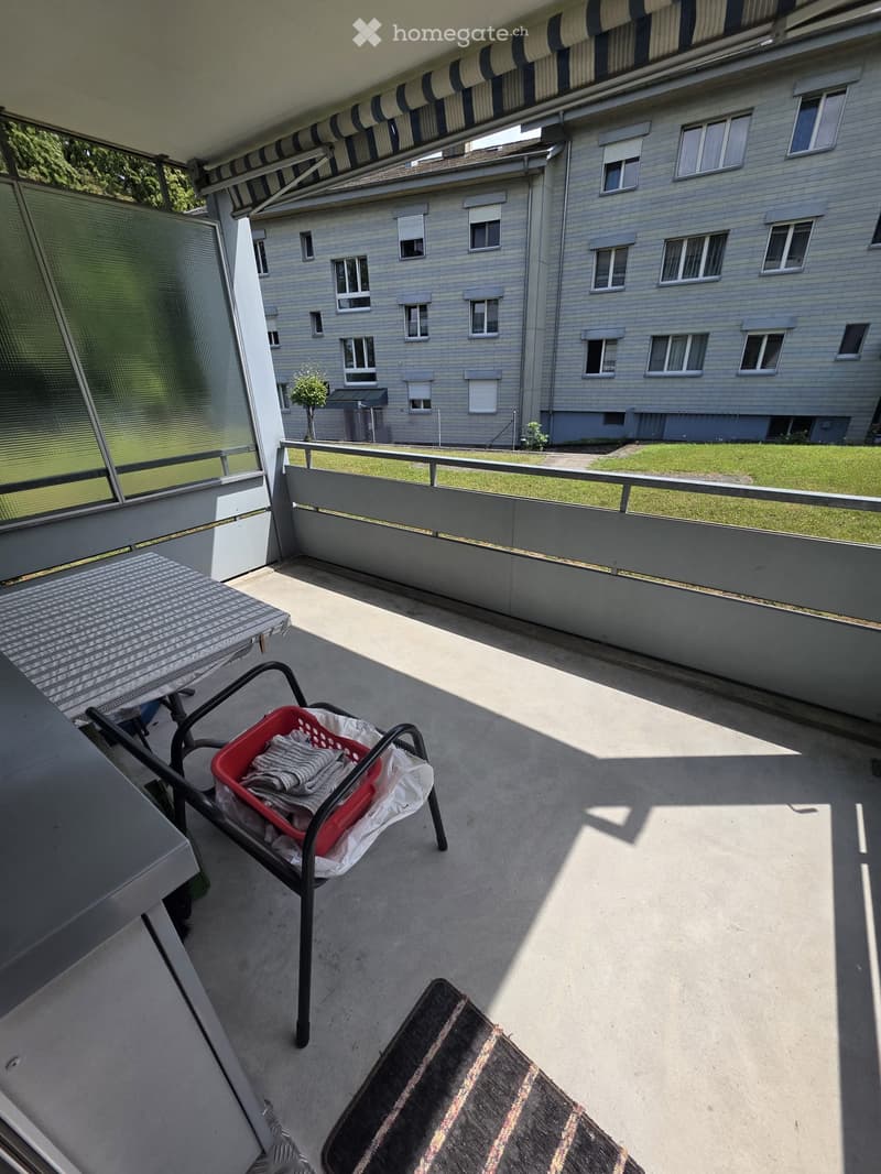 Balcony with a table, chair, laundry basket, and rug, overlooking a grassy area and a multi-story building.