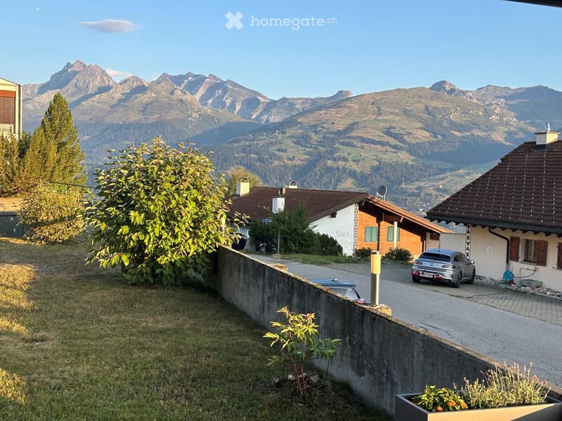 A house with a brown tiled roof and white walls, situated in a scenic valley with mountains in the background. There is a driveway with a parked car and some greenery in the foreground.