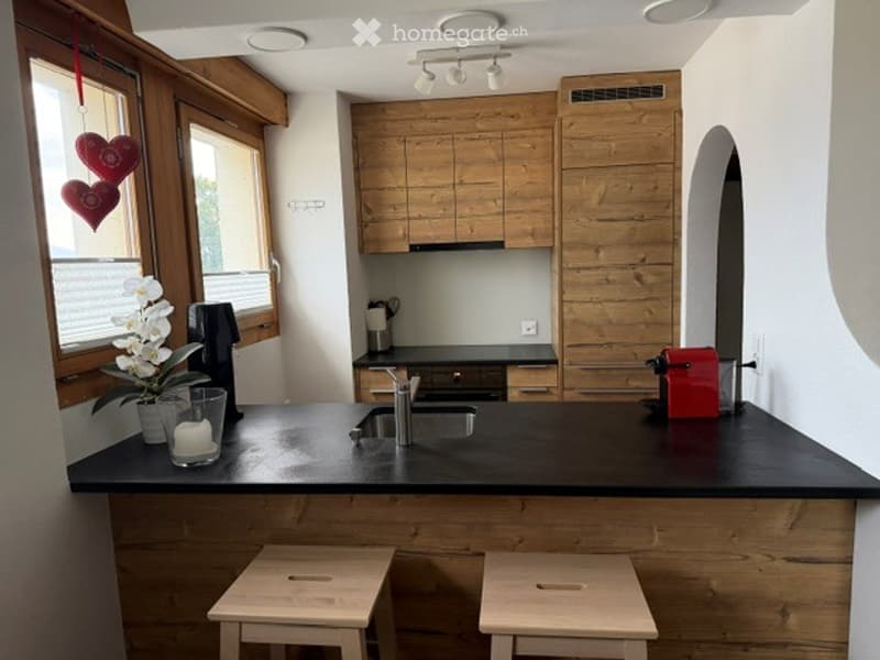 Modern kitchen with wooden cabinets, black countertop, sink, and two wooden stools. Features a red coffee maker, a candle, and a vase with white flowers on the counter.