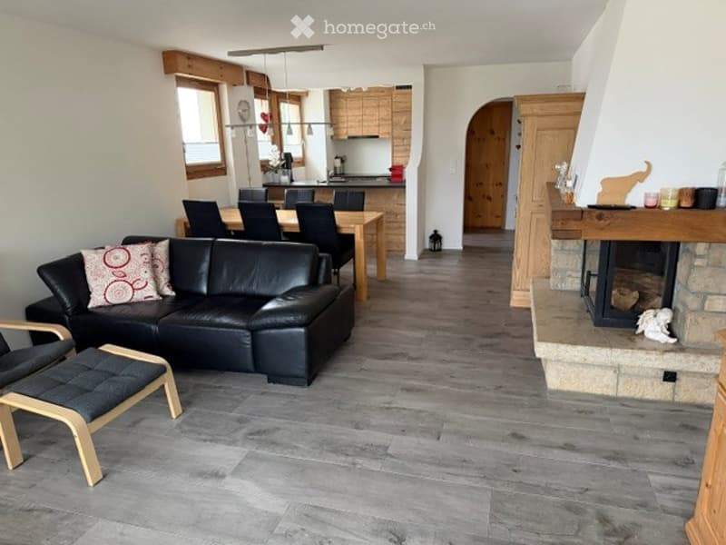 Open-plan living and dining area with grey leather sofa, coffee table, armchairs, ottoman, dining table with chairs, and a fireplace. Kitchen area with wooden cabinets and appliances visible.