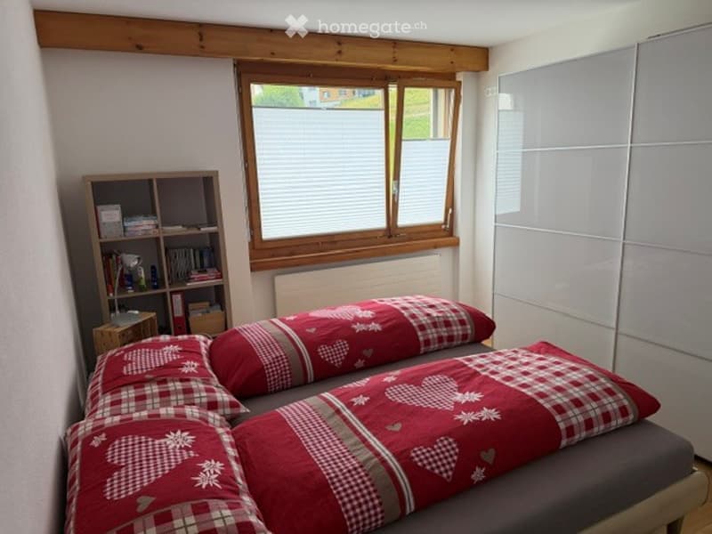 Cozy bedroom with a double bed, red and grey checkered bedding, a wooden nightstand with a lamp, and a bookshelf filled with books. The room features wooden beams on the ceiling and large windows with blinds.