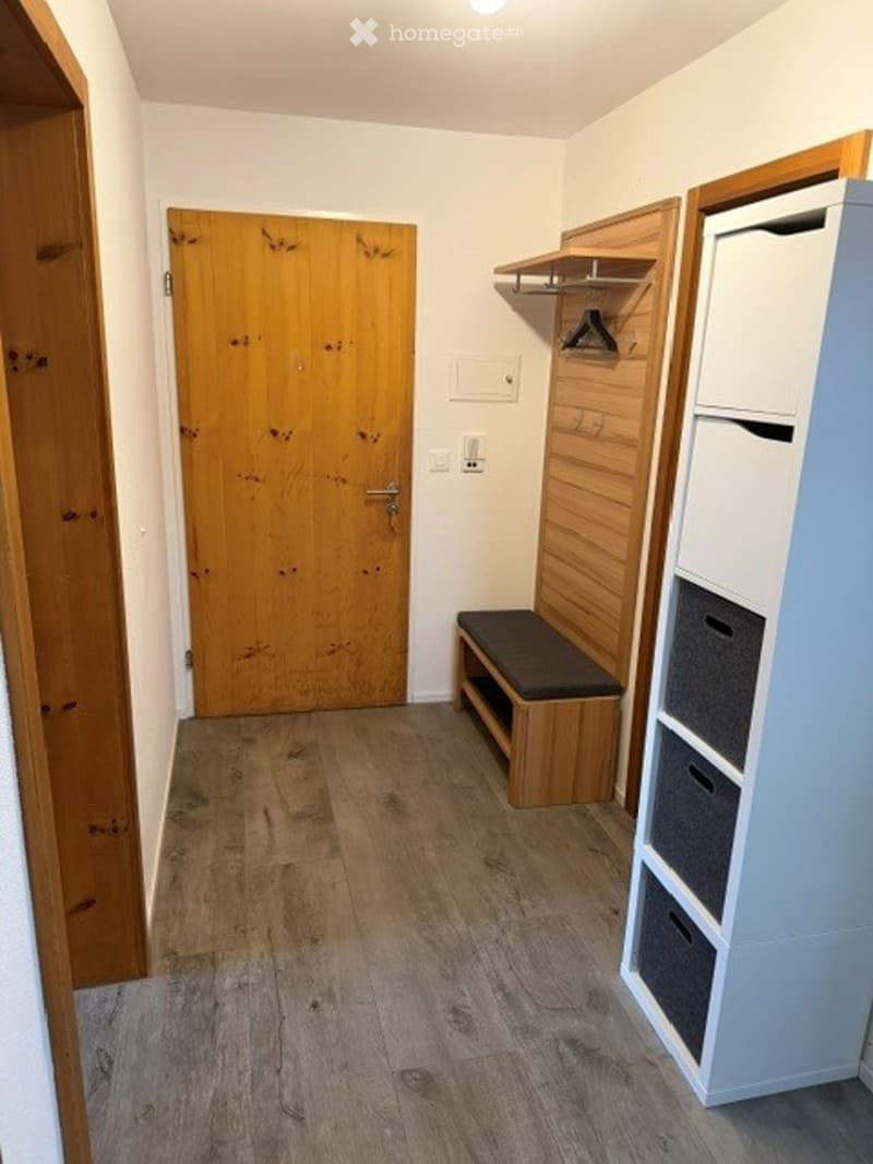 A narrow hallway with wooden flooring, white walls, and a wooden door at the end. There is a small wooden bench with a cushioned seat and a white storage unit with drawers and shelves on the right side.