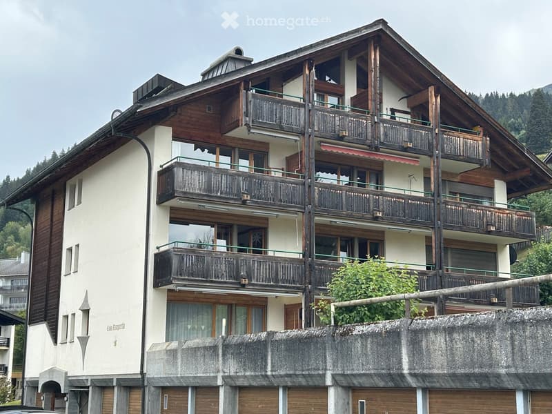 Multi-story residential building with wooden balconies, large windows, and a sloped roof, situated in a mountainous area.