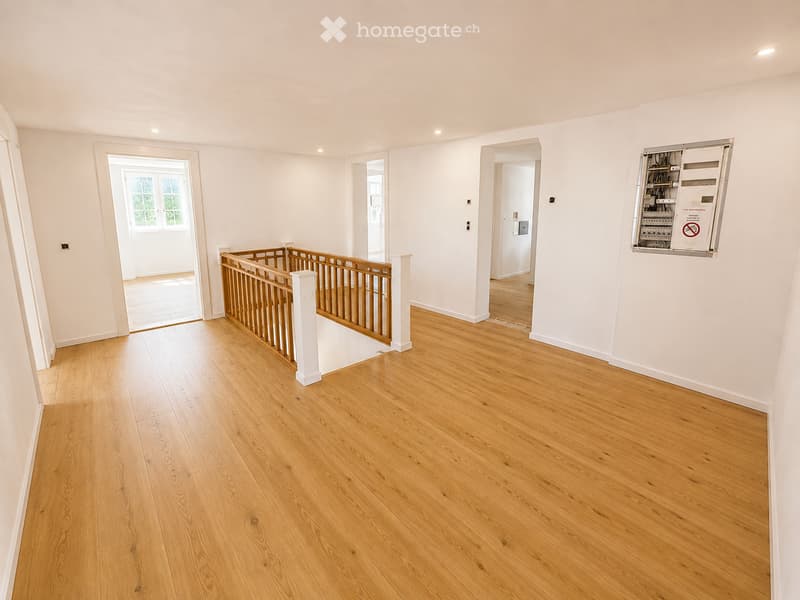 Empty hallway with wooden flooring, white walls, and a staircase with a wooden railing. There are several doorways leading to other rooms.