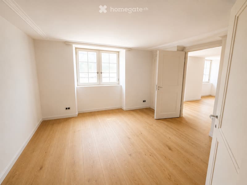 Empty room with light wooden flooring, white walls, and a large window with multiple panes. A door leads to another room with similar flooring.