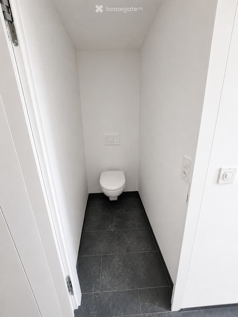 Modern, compact bathroom with white walls, black tiled floor, and a single toilet. White electrical outlets and switches are visible on the walls.
