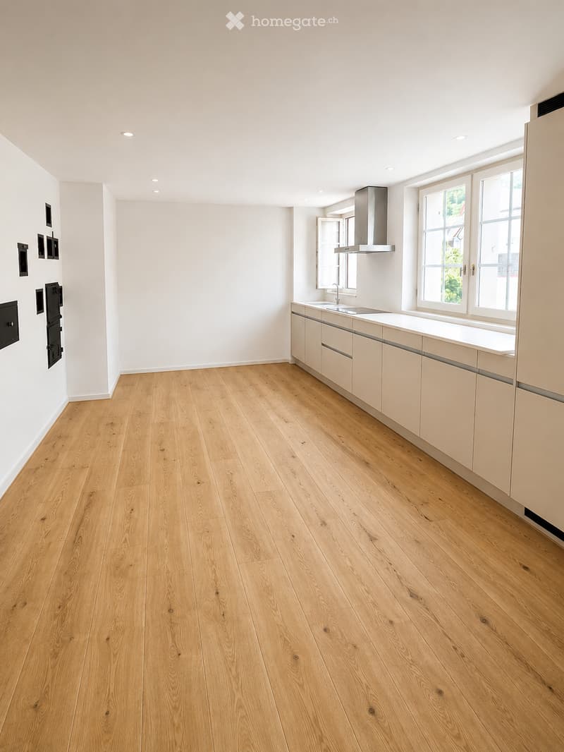 Empty kitchen with light wooden flooring, white cabinetry, and a stainless steel range hood. The space is well-lit with recessed ceiling lights and large windows.