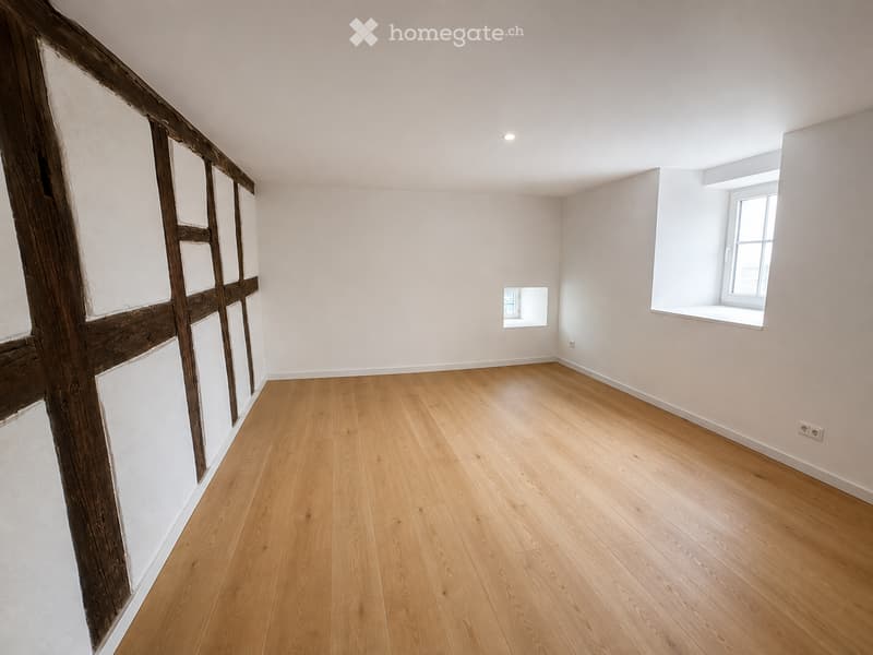 Empty room with light wooden flooring, white walls, and two windows allowing natural light. Exposed wooden beams on one wall.