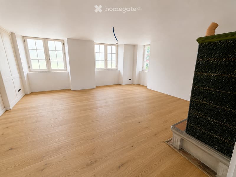 Empty room with light wooden flooring, white walls, and multiple windows allowing natural light. A decorative fireplace is visible on the right side.