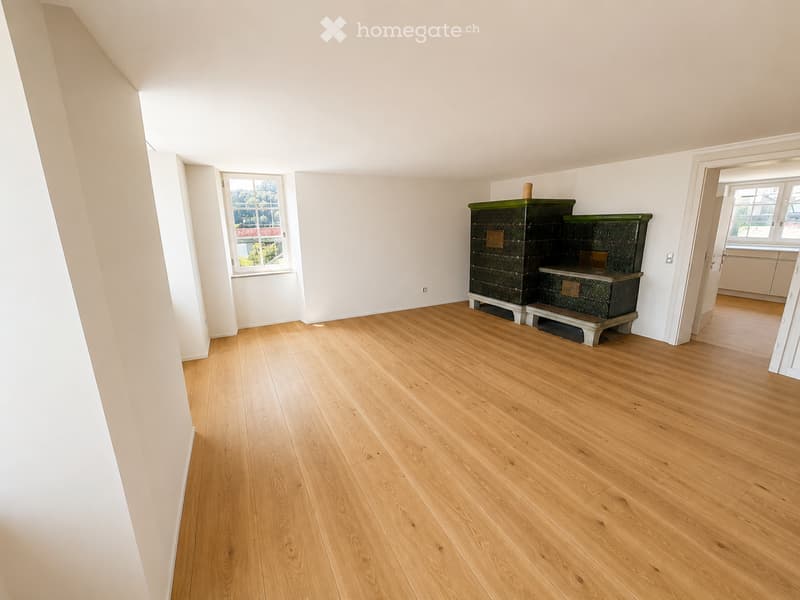 Empty room with light wooden flooring, white walls, and a fireplace with green tiles. There are windows on two walls, and the room is well-lit with natural light.