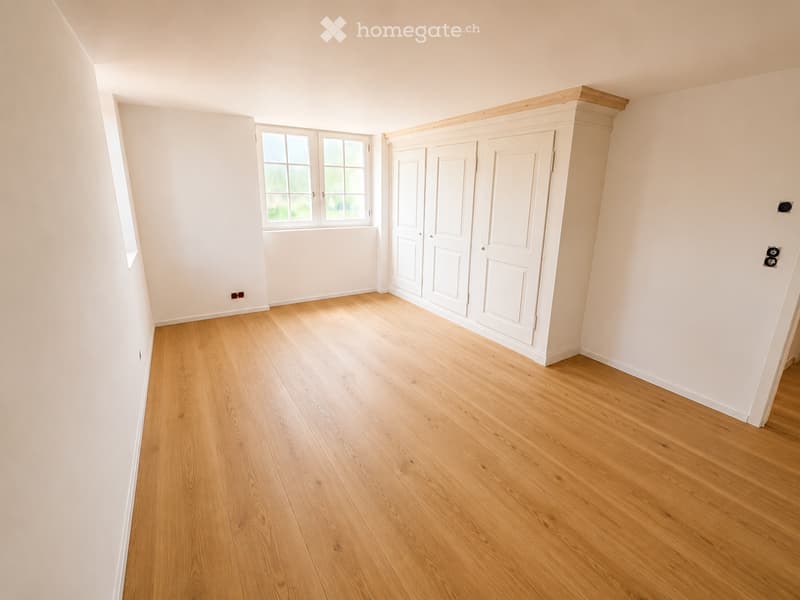 Empty room with light wooden flooring, white walls, and a large window with multiple panes. There is a built-in white cabinet with double doors on one wall.