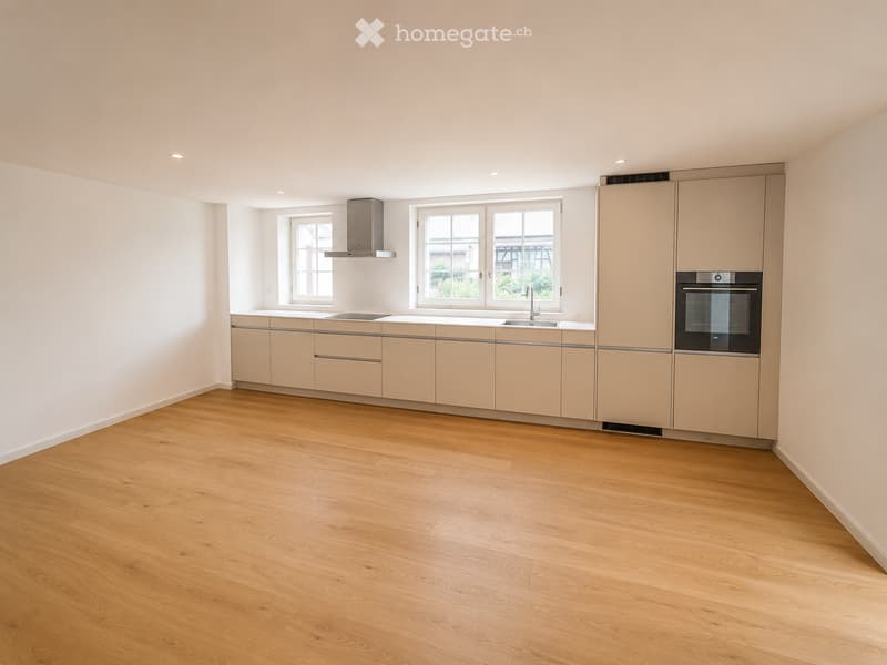 Modern kitchen with light-colored cabinets, wooden flooring, and stainless steel appliances including a sink, faucet, and oven. Natural light from windows illuminates the space.