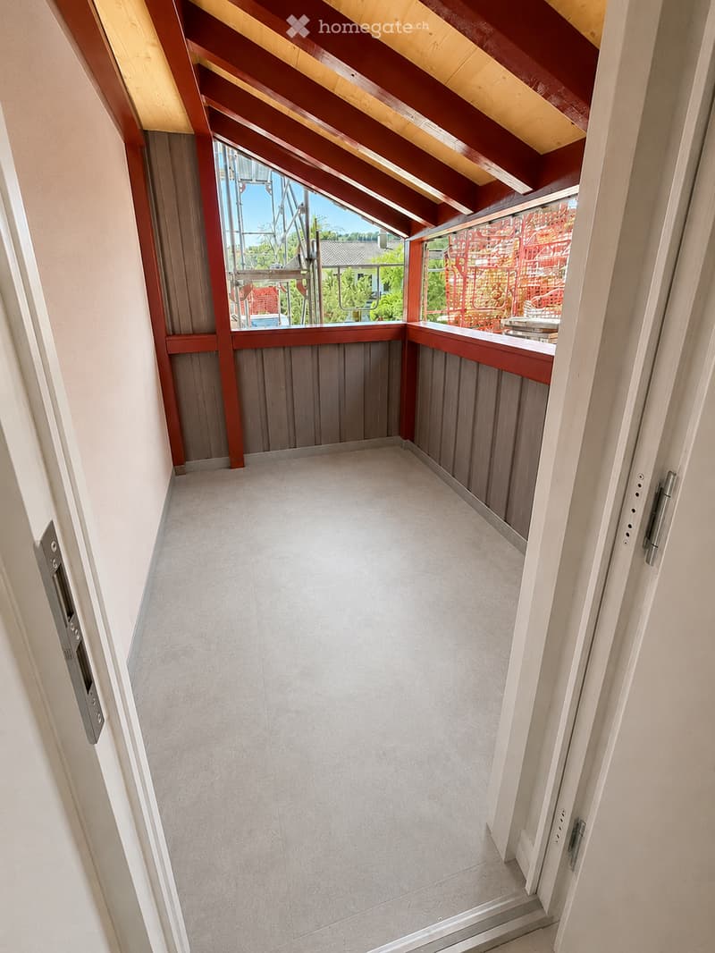 Balcony with wooden ceiling beams, grey tiled floor, and grey wooden paneling on the walls. The balcony has large windows offering a view of the outdoor area.