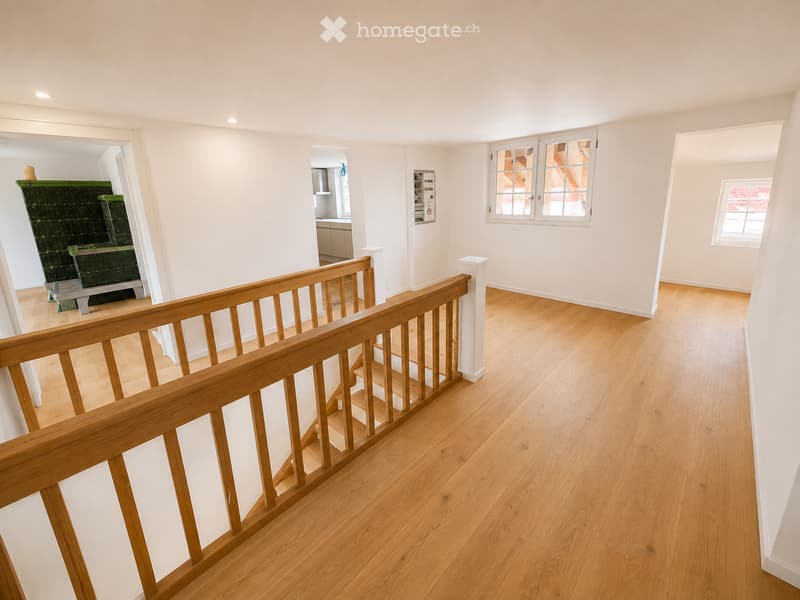 Bright, open hallway with wooden flooring and a staircase leading upwards, featuring white walls and large windows allowing natural light to fill the space.
