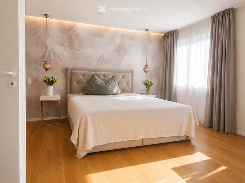 Elegant bedroom with a tufted headboard, white bedding, wooden floor, and two pendant lights. Two nightstands with vases of yellow flowers flank the bed.