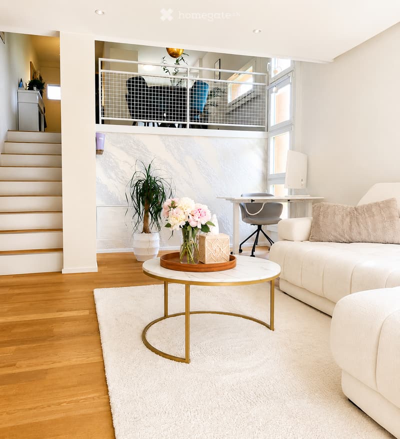 Modern living room with white walls, wooden flooring, a white sofa, a round coffee table with a gold frame, a white shag rug, a potted plant, and a vase with flowers on the coffee table. A staircase leads to an upper level with a railing.