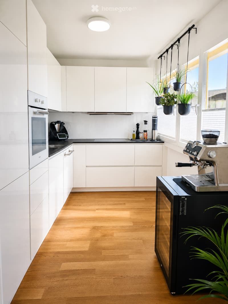Modern kitchen with white cabinets, wooden flooring, and stainless steel appliances including an oven, coffee machine, and refrigerator. Potted plants are visible on the windowsill and counter.