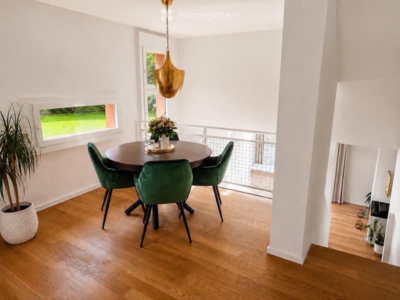 Dining area with a round wooden table, four green velvet chairs, a hanging gold pendant light, and a potted plant near the window.