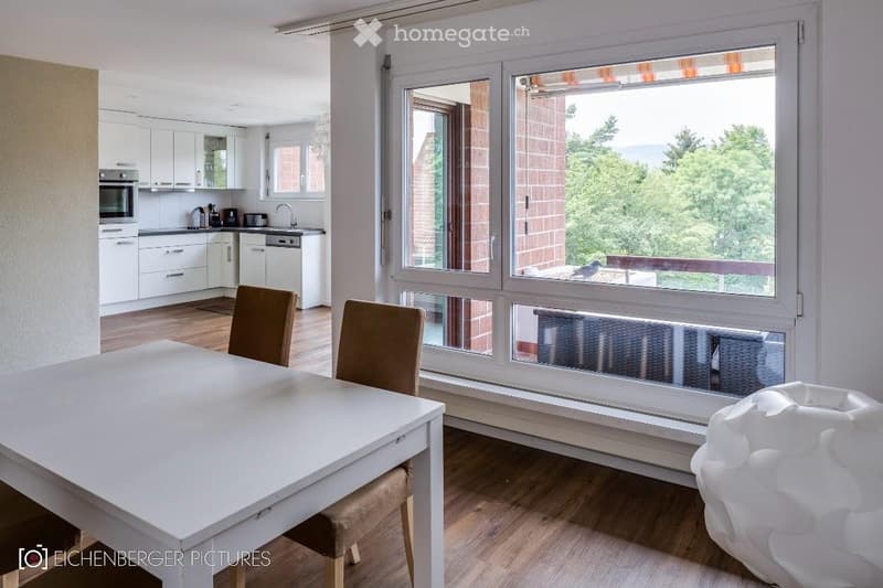 Bright, open-plan dining and kitchen area with large windows offering a scenic view. The dining area features a white table with two chairs, while the kitchen includes white cabinets, an oven, a dishwasher, and a sink.