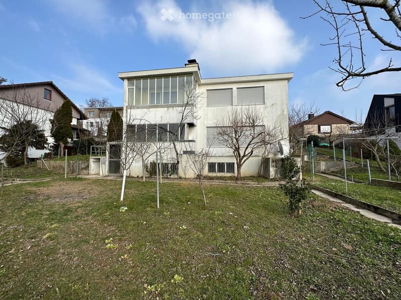 Modern two-story building with large windows and a flat roof, surrounded by a grassy yard with sparse trees and neighboring houses.