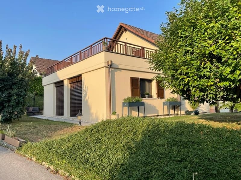Two-story house with beige exterior, brown shutters, and a balcony on the upper floor. The front yard features a well-maintained lawn, small garden beds, and outdoor furniture.