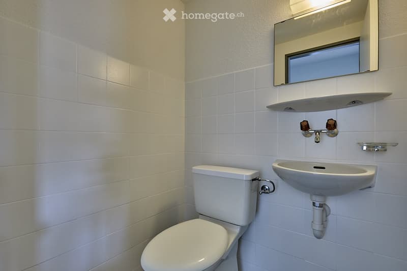 white tiled walls, white ceramic sink, silver faucet, white ceramic toilet, small mirror with window