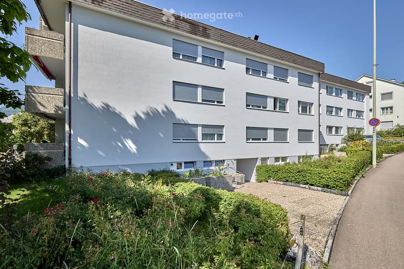 Apartment building with white exterior, multiple balconies, windows with shutters, small gardens in front