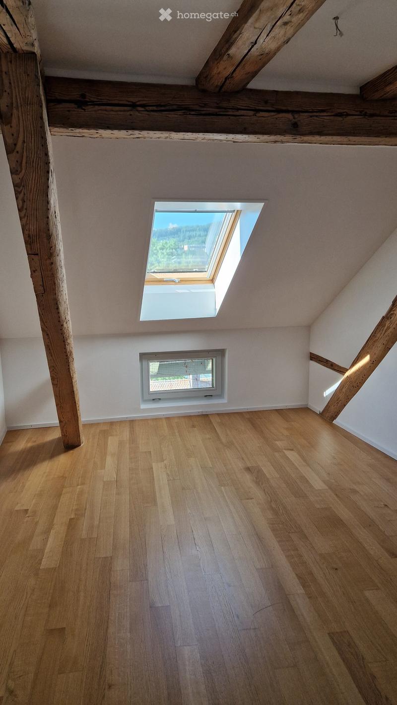 Empty attic room with wooden floors, exposed wooden beams, and two windows, one of which is a skylight offering a view of the outdoors.