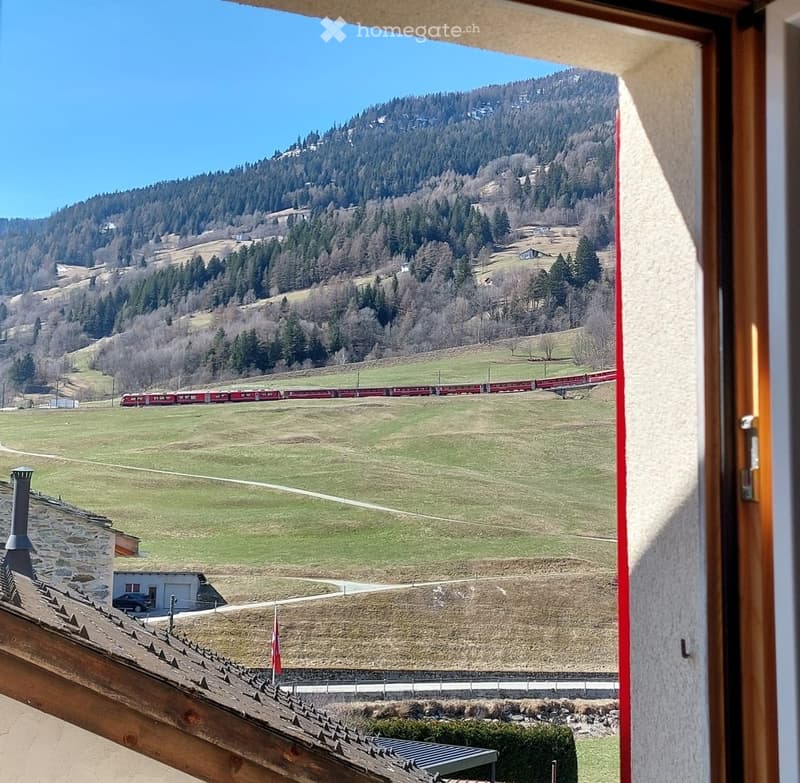 View from a balcony showing a scenic landscape with a train passing through a valley, surrounded by green fields and forested hills.