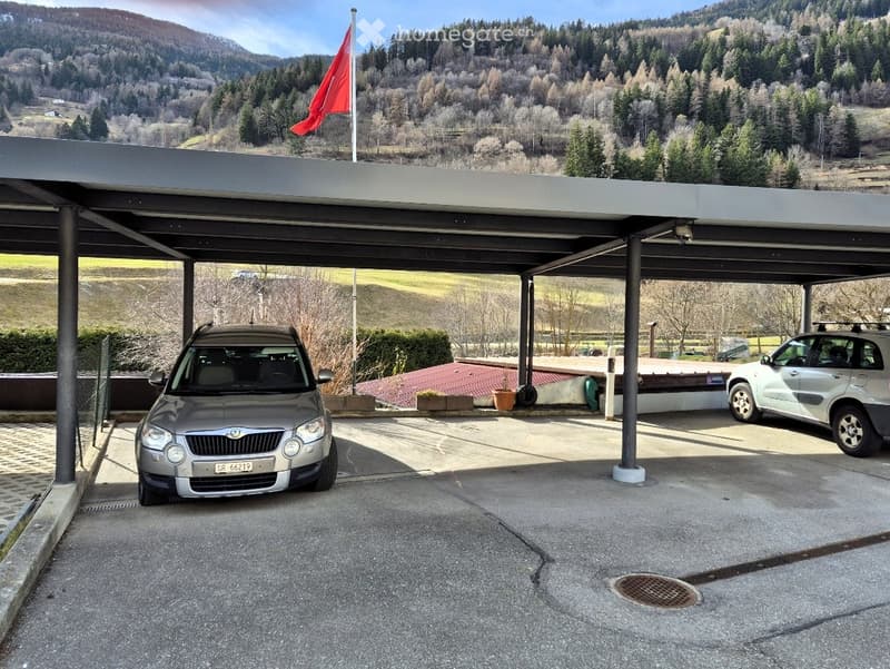 Carport with two parked cars, surrounded by a scenic mountainous landscape with trees and a flagpole.