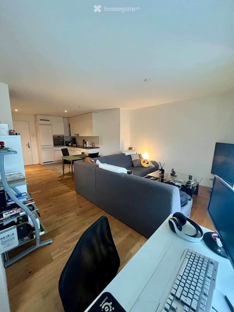 A modern living room with a grey sofa, coffee table, and a small kitchen area in the background. There is a desk with a laptop, keyboard, and headphones, and a chair in front of it.