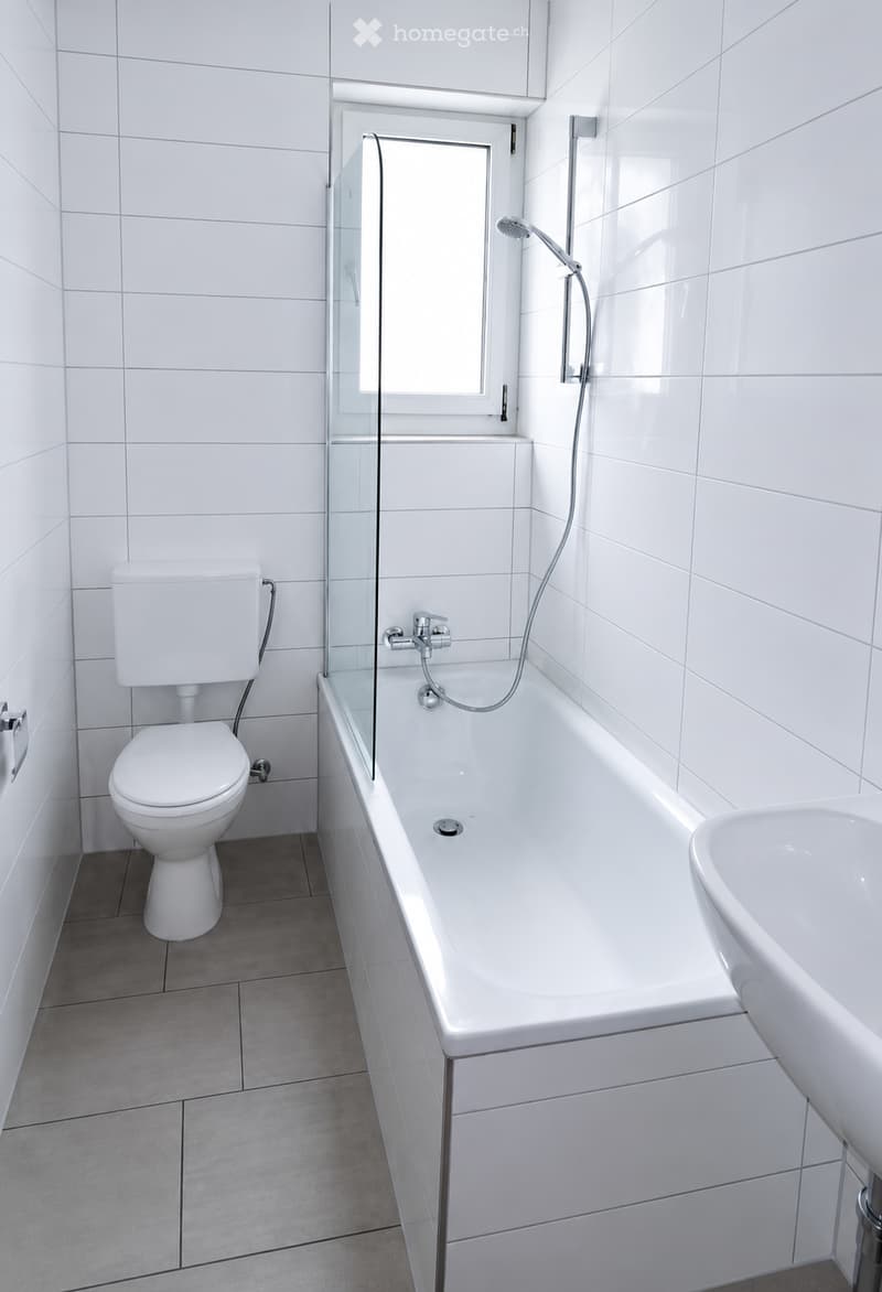 Modern bathroom with white tiles, a bathtub with a glass shower enclosure, a toilet, and a sink.