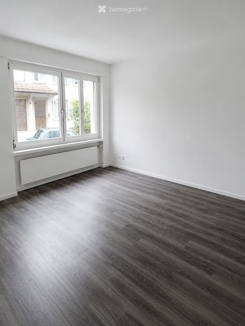 Empty room with dark wooden flooring, white walls, large window with radiator below, and visible electrical outlets.