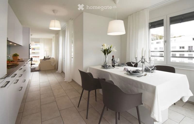 Modern open-plan living and dining area with white walls, tiled floor, and large windows. Features a dining table with chairs, a sofa, kitchen cabinets, and decorative items.