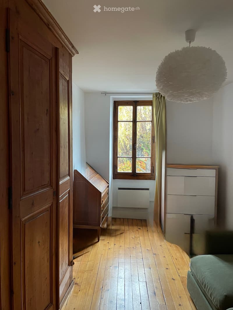 Bright bedroom with wooden flooring, large window with wooden frame, green bed, wooden wardrobe, and a hanging pendant light.
