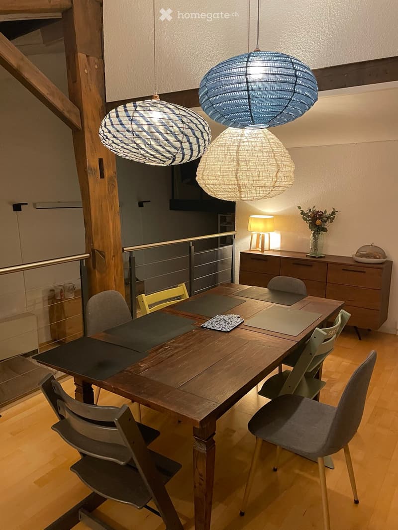 Dining area with a wooden table, multiple chairs, pendant lights, a side table with a lamp, and a vase with flowers.