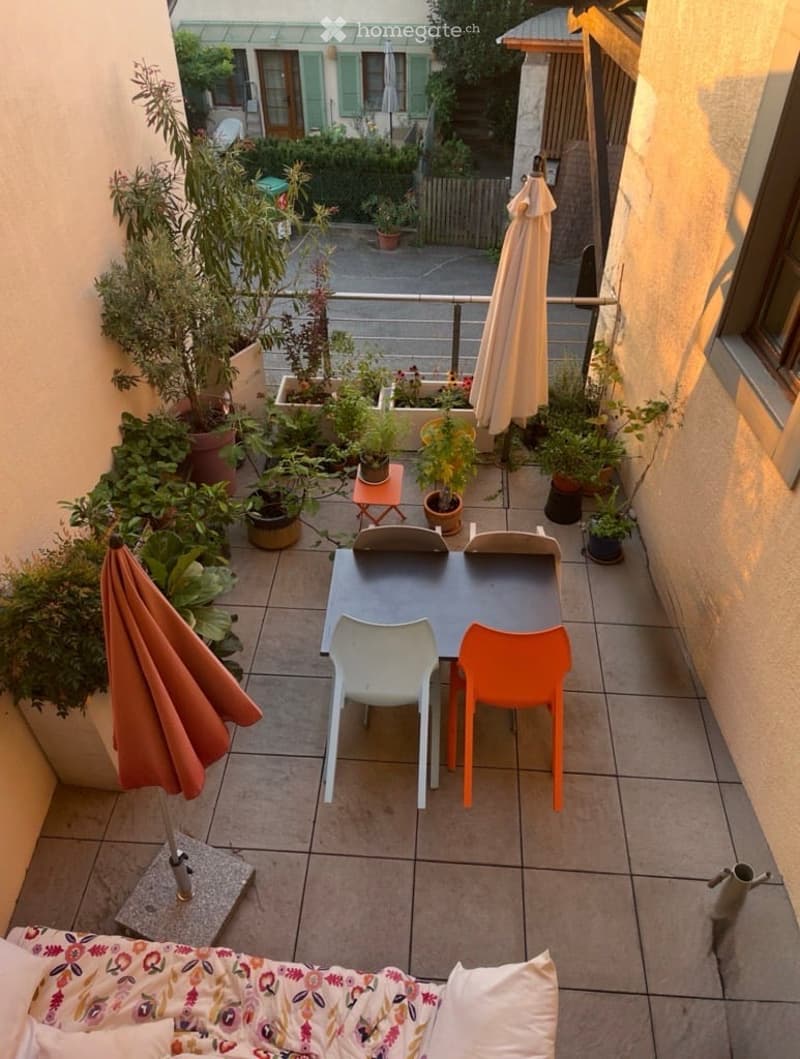 Balcony with tiled floor, outdoor furniture including a table, chairs, and potted plants, with a view of a garden and building in the background.