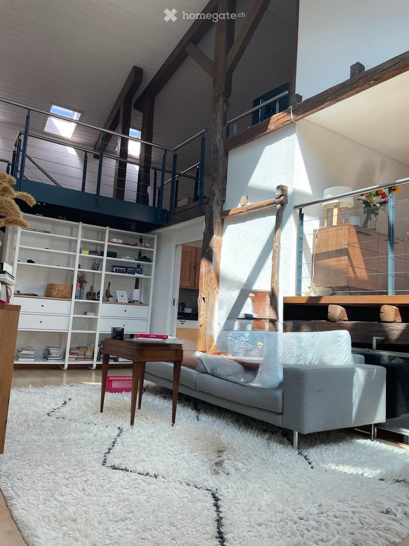 Modern living room with a grey sofa, coffee table, bookshelf, and a large white shag rug. The room features exposed wooden beams and a staircase leading to an upper level.