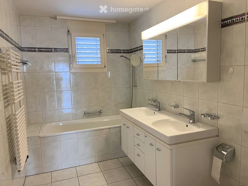 Modern bathroom with a bathtub, double sink, large mirror with built-in lighting, and a radiator. The walls are tiled in beige with decorative border tiles.