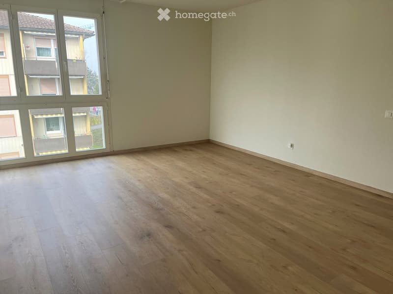 Empty room with wooden flooring and large windows offering a view of neighboring buildings and balconies.