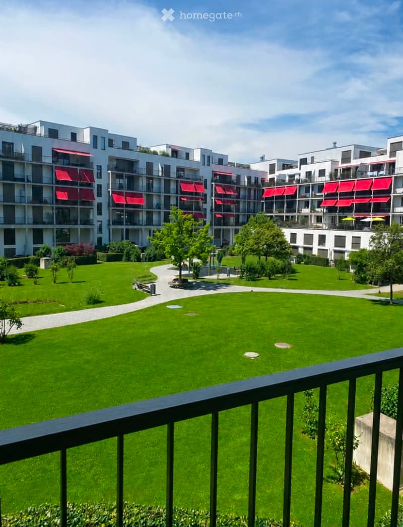 View from a balcony overlooking a well-maintained green lawn and modern residential buildings with red awnings.