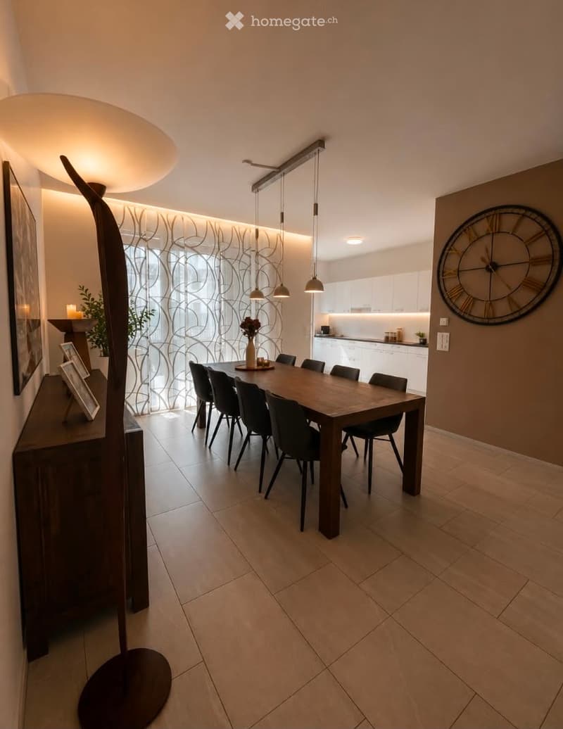 Modern dining area with a wooden dining table surrounded by black chairs, a floor lamp, a cabinet with picture frames, and a large wall clock. The space features tiled flooring and large windows with decorative curtains.