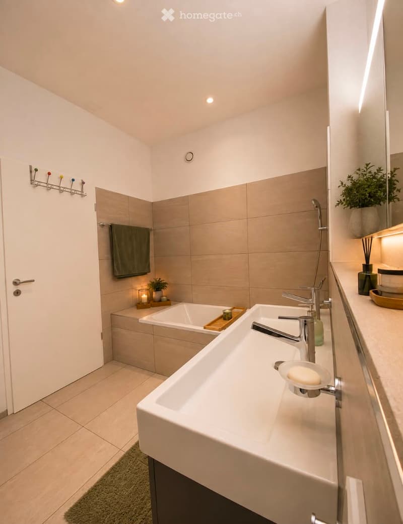 Modern bathroom with a large sink, bathtub, and a mirror. Features include a towel rack, a candle, and decorative plants.