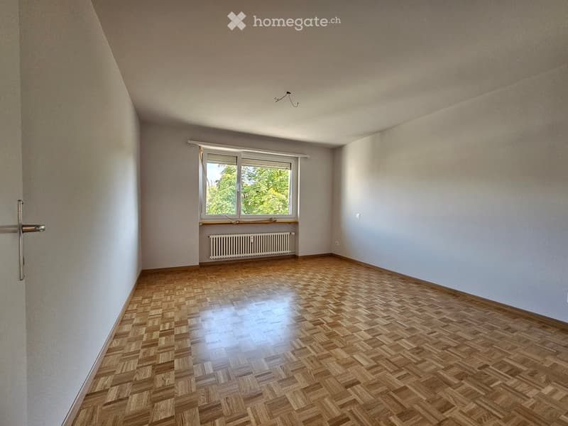 Empty room with wooden parquet flooring, white walls, and a large window with a radiator below it.